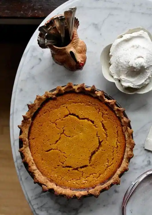 A pumpkin pie sitting on a marble table next to a turkey cup holding spoons and a bowl of whipped cream. 