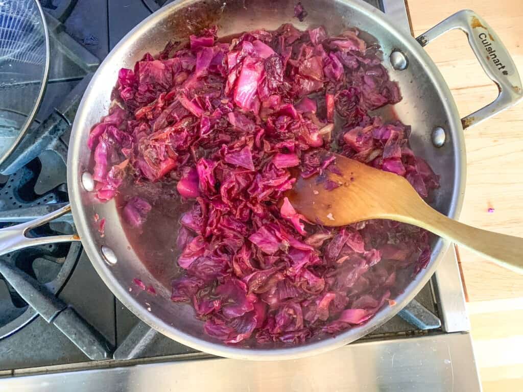 Sweet and sour cabbage cooking in a frying pan on the stove with a wood spoon in it. 