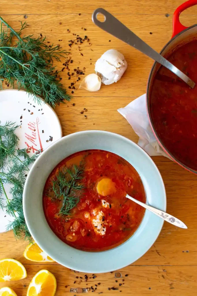 A blue bowl filled with tomato rice soup on a wood table next to a pot of soup and some garlic and red pepper flakes with dill. 