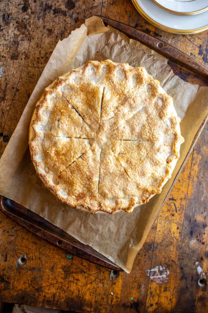 An entire apple pie sitting on a baking sheet that is covered with parchment paper sitting on a wood table. 