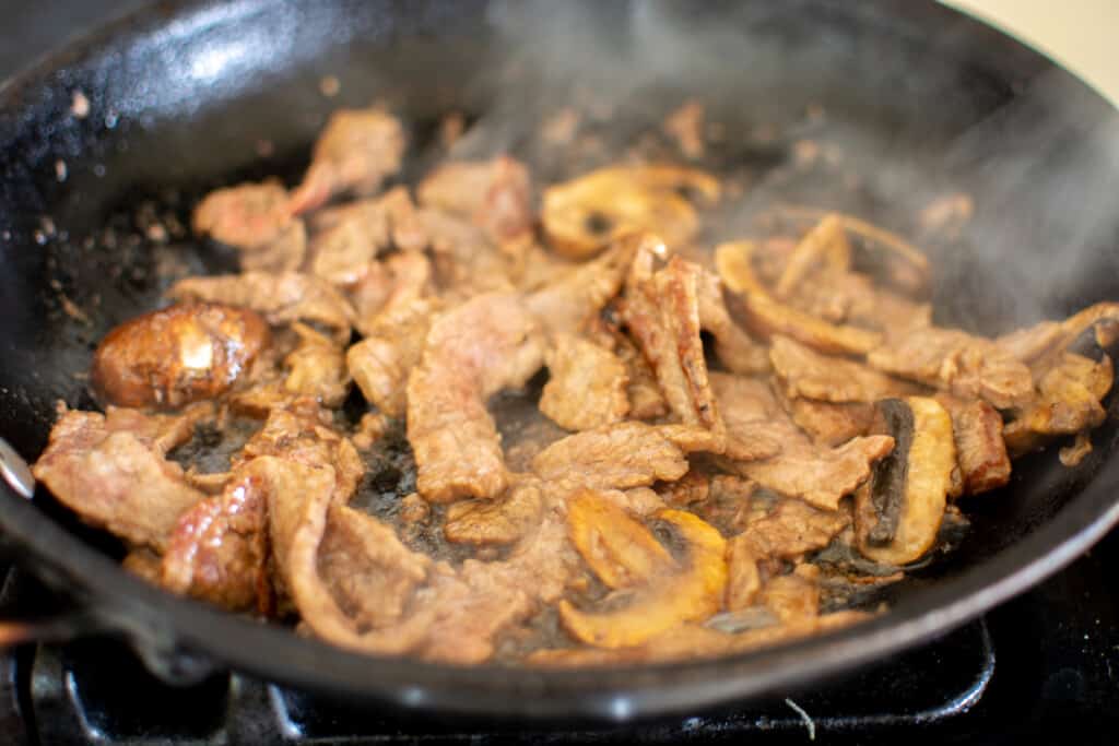 A black frying pan on the stove filled with strips of beef and sliced mushrooms sautéing in oil. 