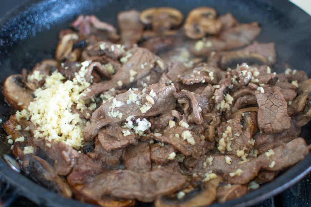 A black frying pan on the stove filled with strips of steak and sliced mushrooms with chopped garlic and ginger. 