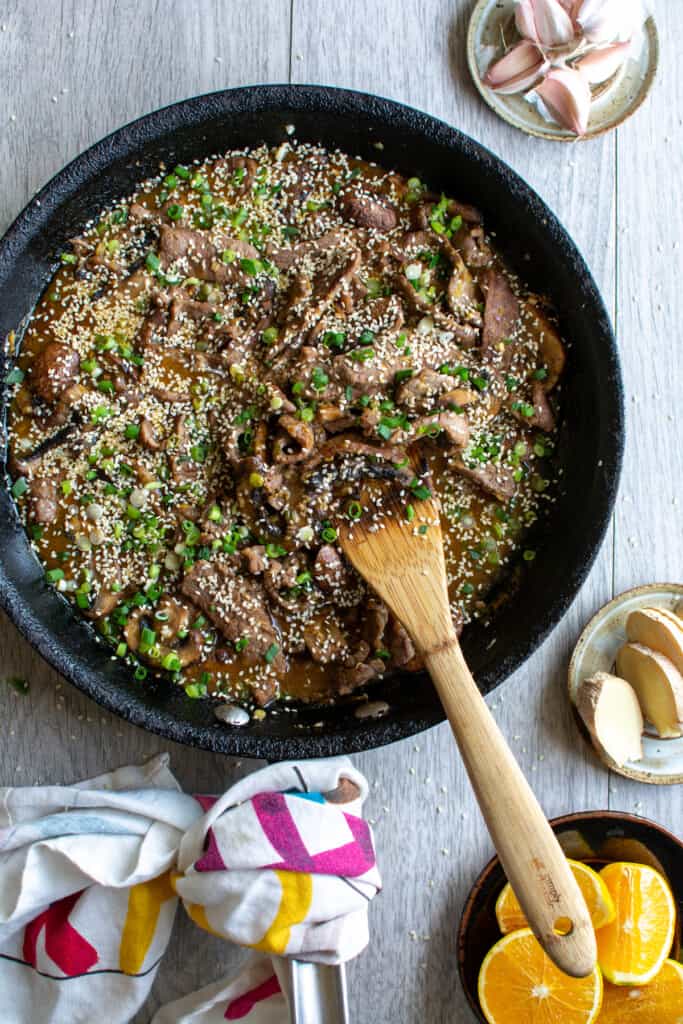 A black frying pan filled with beef stir fry sitting on a gray table next to a little plate of orange wedges.