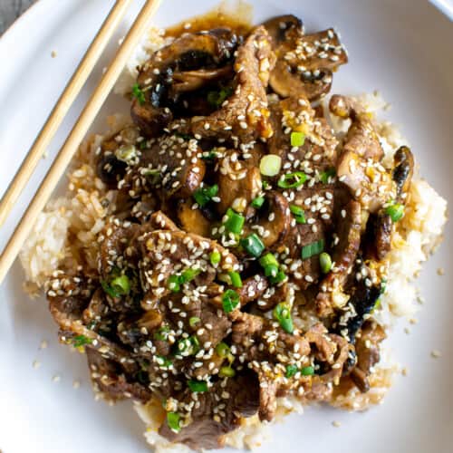 A white bowl filled with rice and beef stir fry topped with sesame seeds and scallions with a pair of chopsticks on the side.