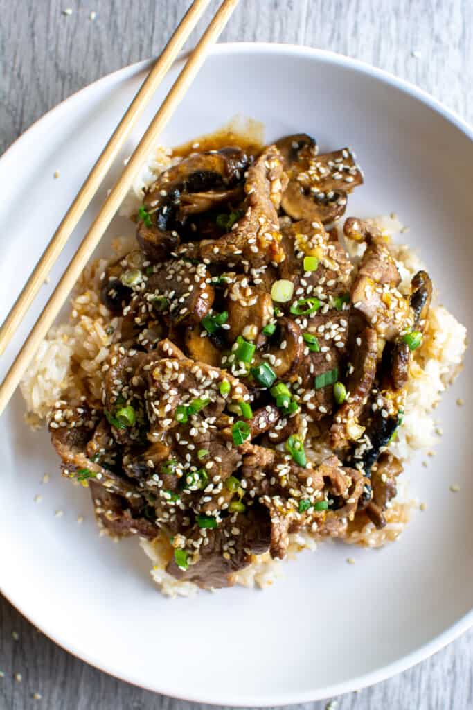 A white bowl filled with rice and beef stir fry topped with sesame seeds and sliced scallions with a pair of chopsticks. 