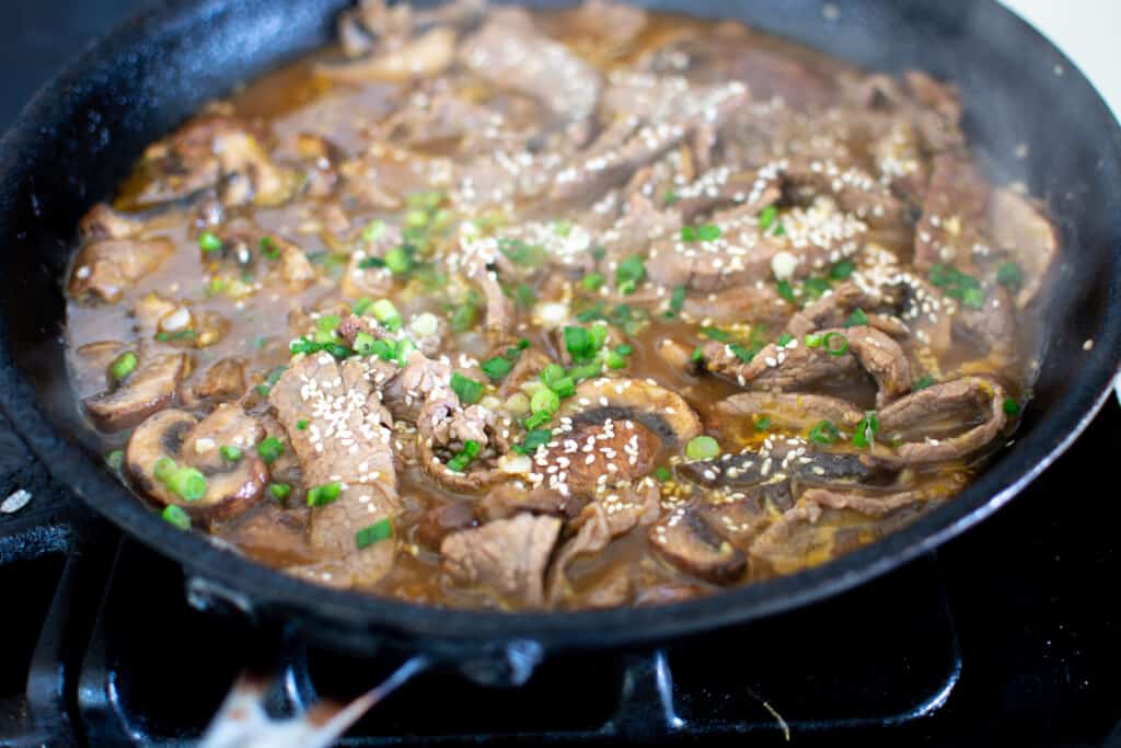 A black frying pan on the stove filled with beef stir fry topped with sliced green onion and sesame seeds and sauce. 