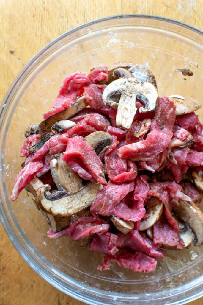 A glass bowl filled with sliced beef and sliced mushrooms that have been tossed in cornstarch sitting on a wood table. 
