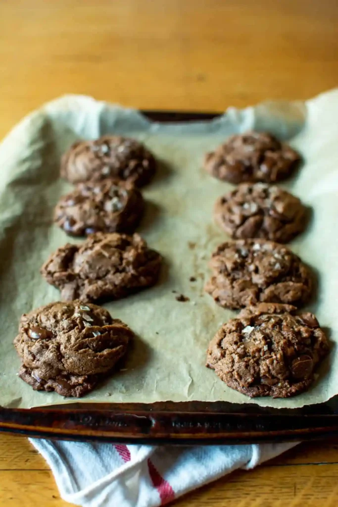 Cocoa cookies sitting on a baking sheet that is covered in parchment paper sitting on a kitchen towel on a wood table. 