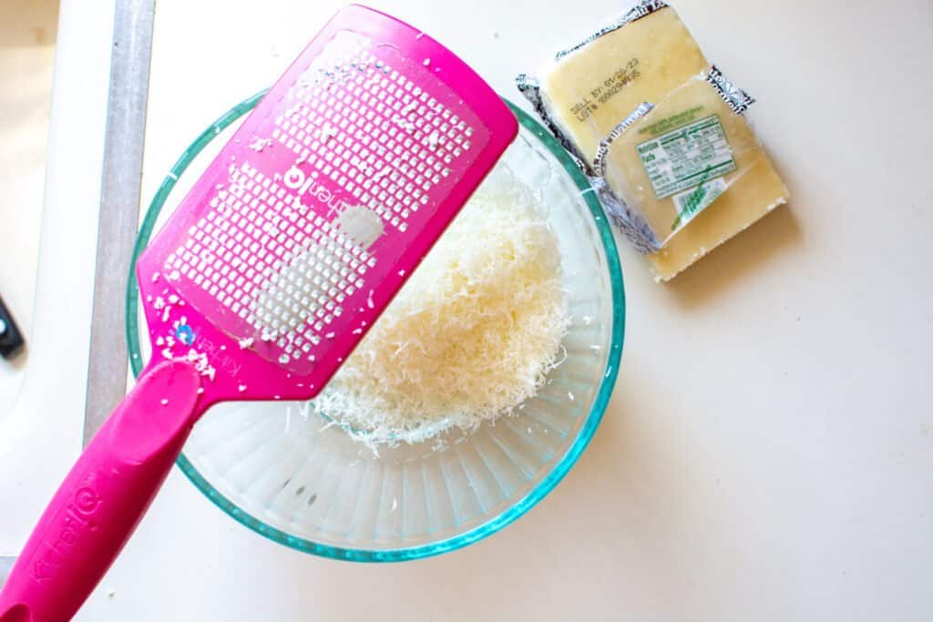 A pink microplane grater sitting over a glass bowl filled with shredded pecorino Romano cheese on a white counter. 