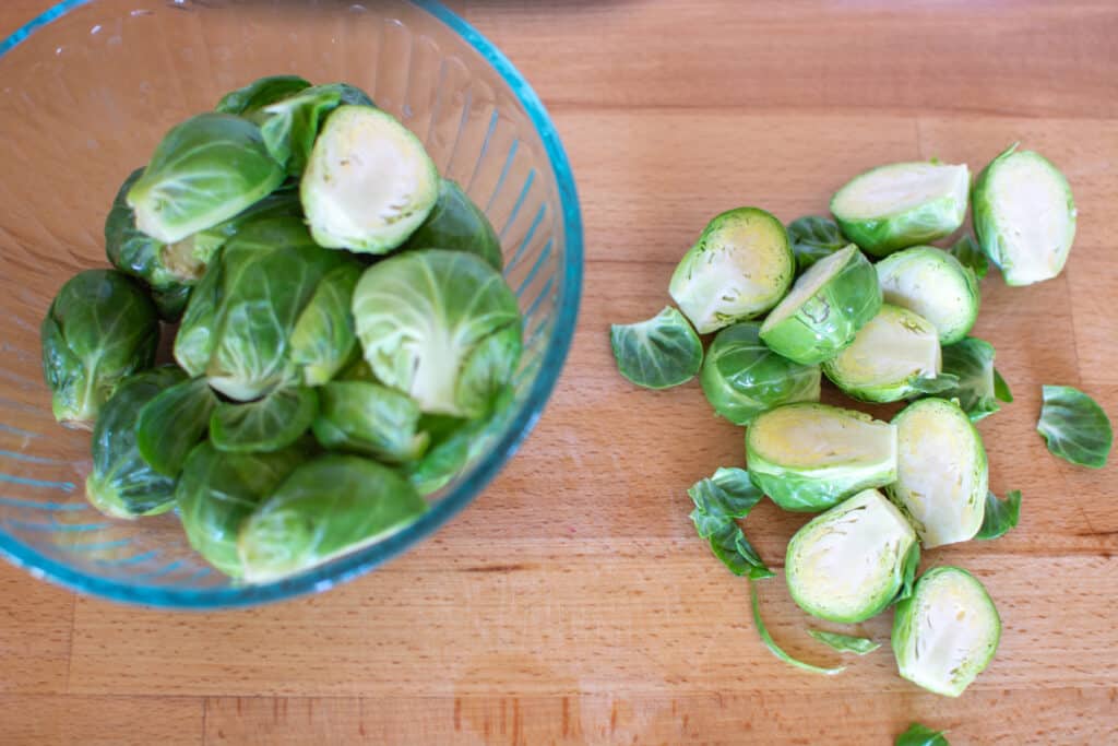 A glass bowl on a wood counter half filled with Brussels sprouts and more Brussels sprouts cut in half on the counter