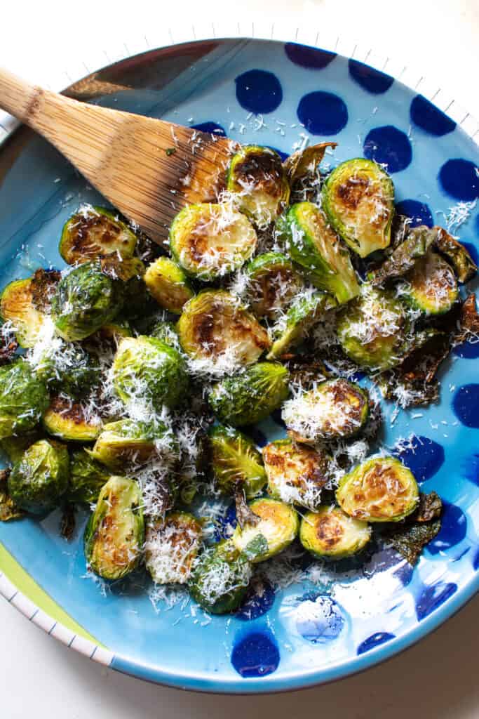 A blue dish with roasted Brussels sprouts on it topped with pecorino cheese with a wood spoon on the side. 