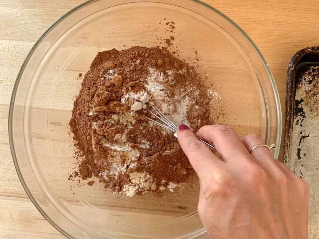 A woman mixing dry ingredients with a whisk in a glass bowl that is sitting on a wood counter. 