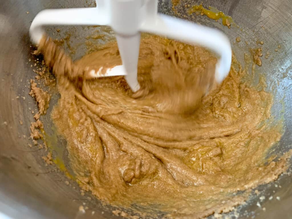 An electric mixer beating vegetable shortening, sugars, and eggs in a metal bowl. 