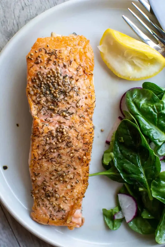 A piece of salmon topped with mustard seed and dill on a white plate with a lemon half, and spinach salad with a fork. 