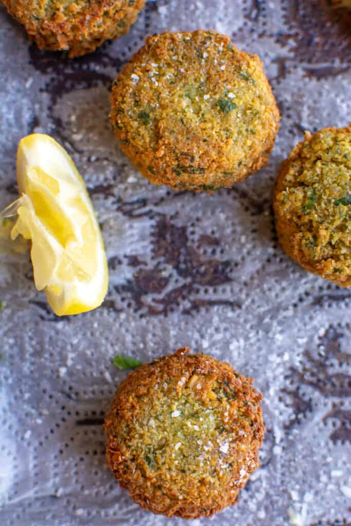 Several fried falafel on a paper towel-lined baking sheet with lemon wedges next to them. 