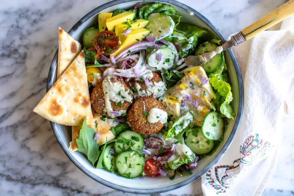 Falafel salad with falafel patties, pita wedges, lettuce, avocado, cherry tomatoes, cucumbers and a fork in the bowl. 