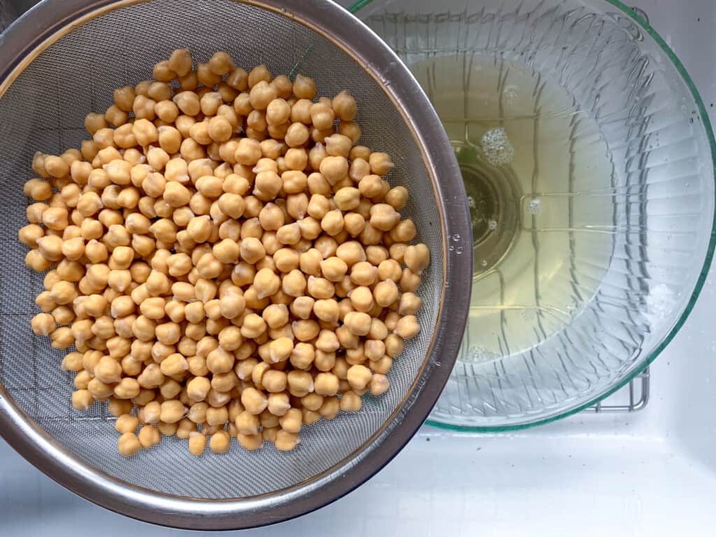 chickpeas that have been soaked in a fine mesh sieve over a large bowl that has the soaking liquid in it. 