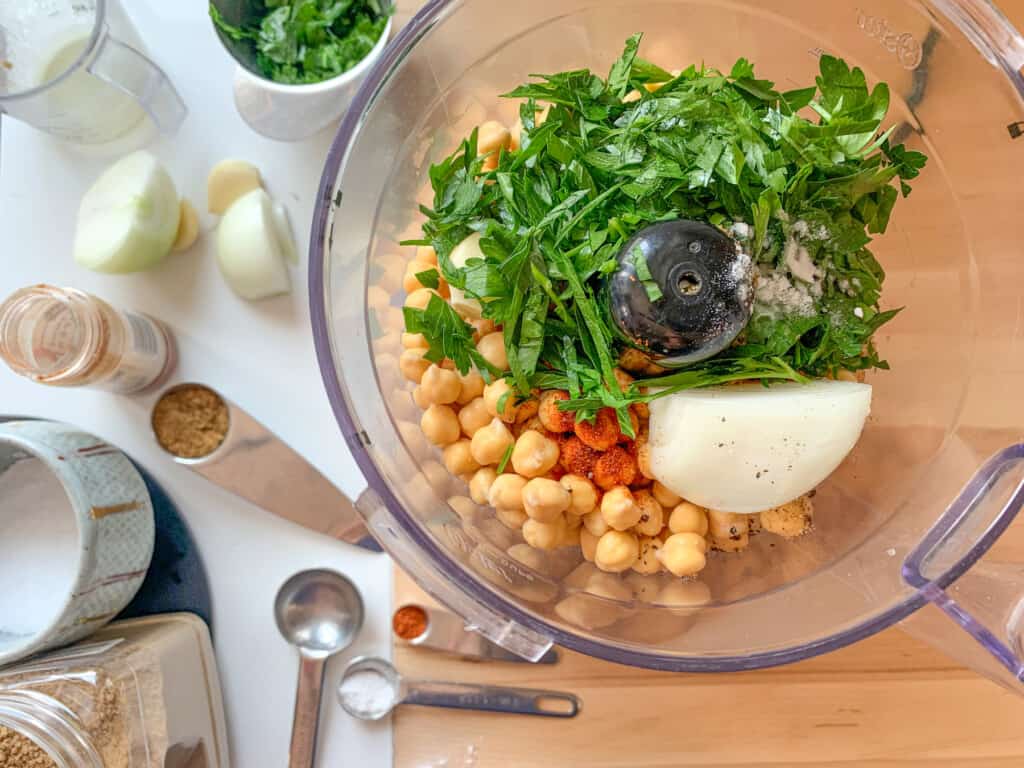 Chickpeas in a food processor topped with parsley, onion, salt, baking soda, cayenne pepper, and garlic with ingredients next to it. 
