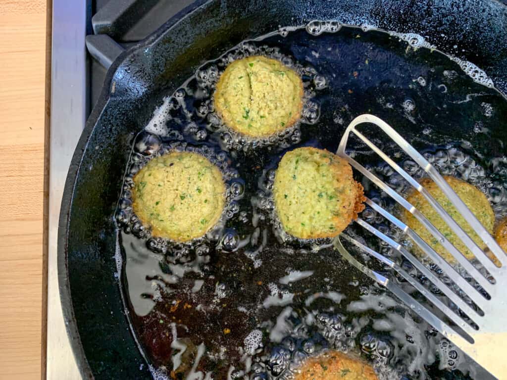 Falafel being fried in a black cast iron skillet with one falafel being flipped with a metal spatula. 