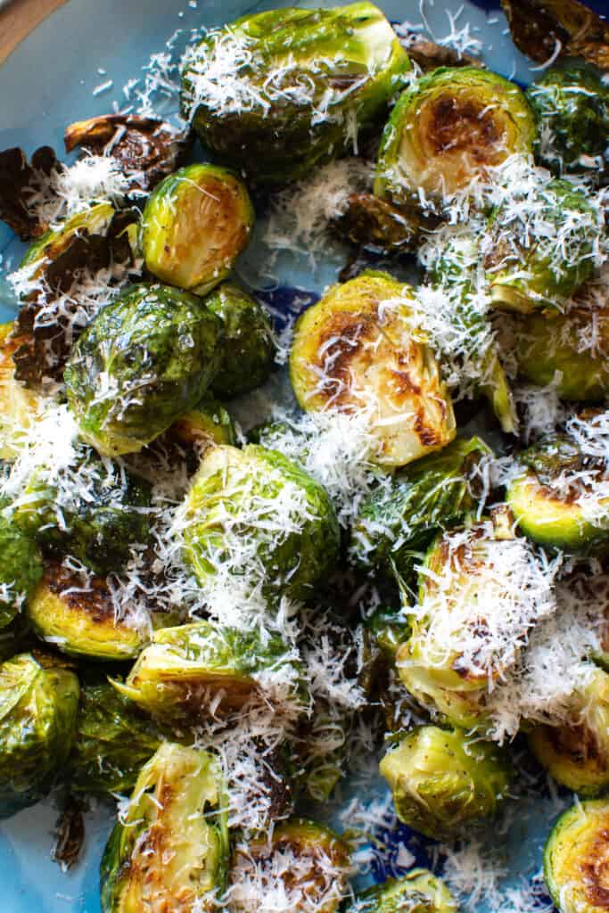 A close up of roasted Brussels sprouts on a blue plate topped with grated pecorino Romano cheese. 