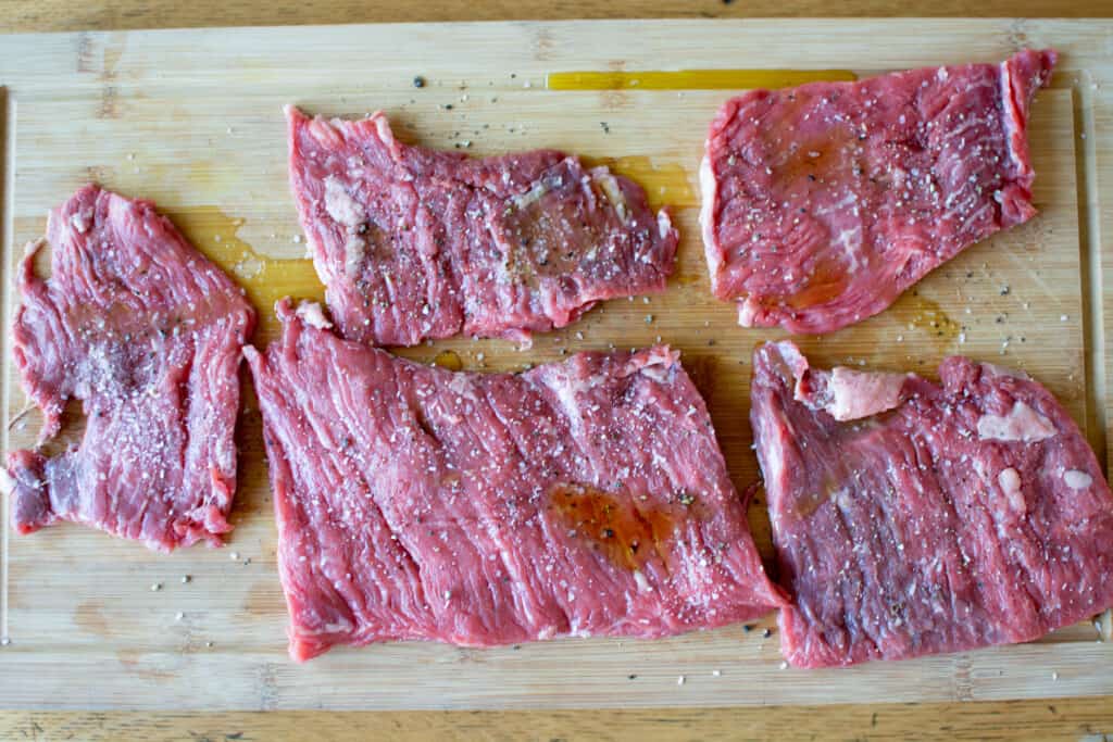 Several pieces of skirt steak on a wood cutting board drizzled with olive oil and sprinkled with salt and pepper. 