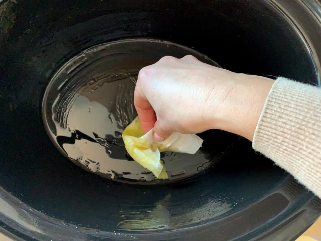 A hand greasing the inside of a crock pot with a paper towel. 