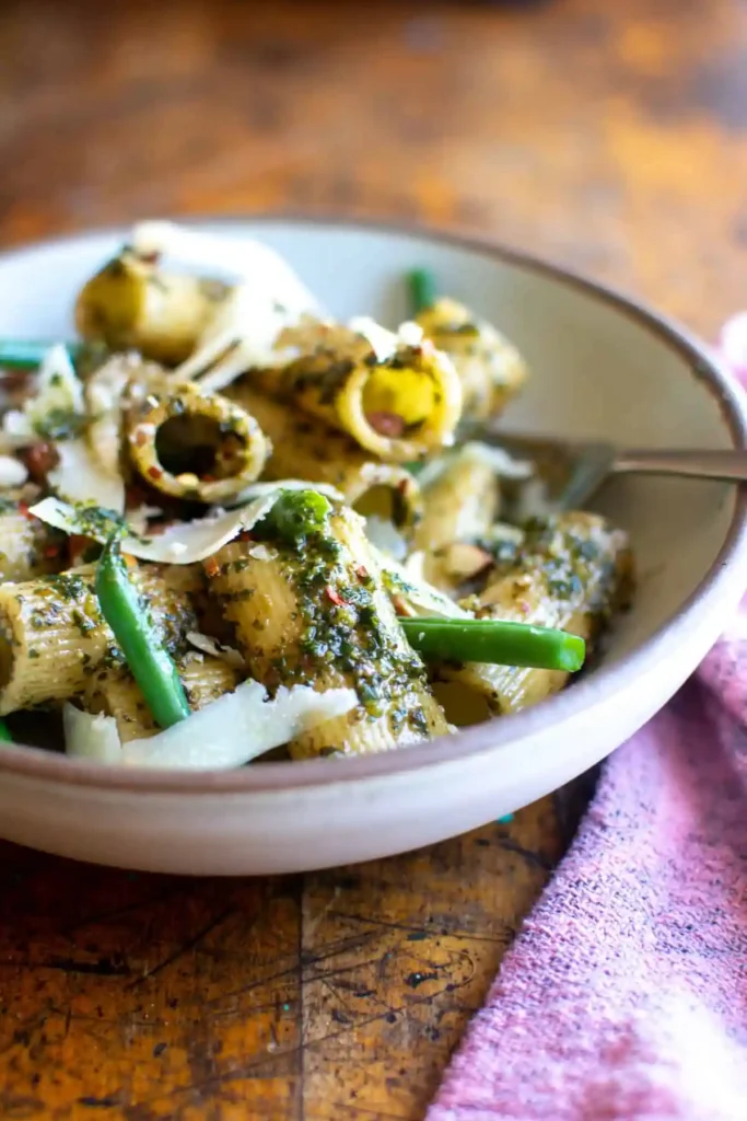 A bowl of vegan pesto pasta on a wood table with a fork sticking out of the bowl and green beans with the pasta. 