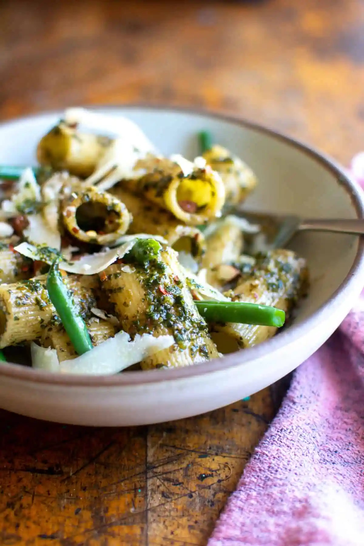 A cream colored bowl filled with rigatoni pasta and green beans with shavings of cheese on top. 