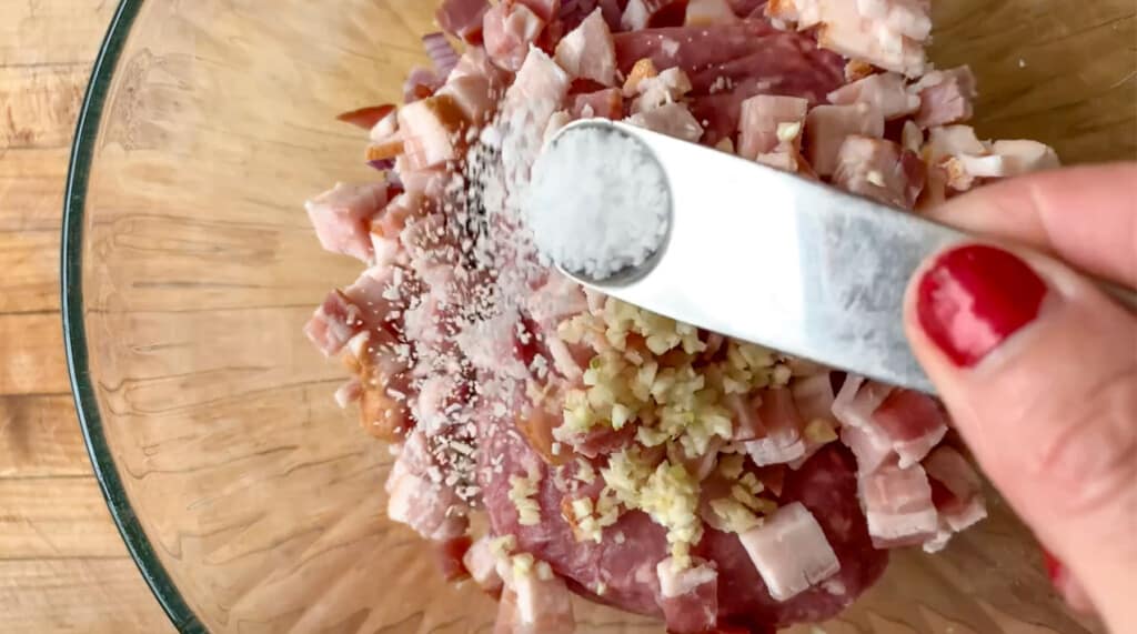 A hand with red finger nails sprinkling salt over a bowl of bacon. 