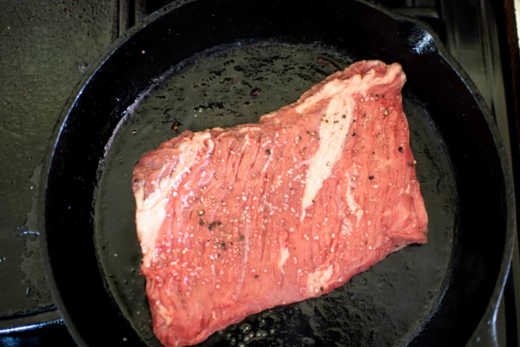A black cast iron pan with a steak cooking in it. The steak is seasoned with salt and pepper. 