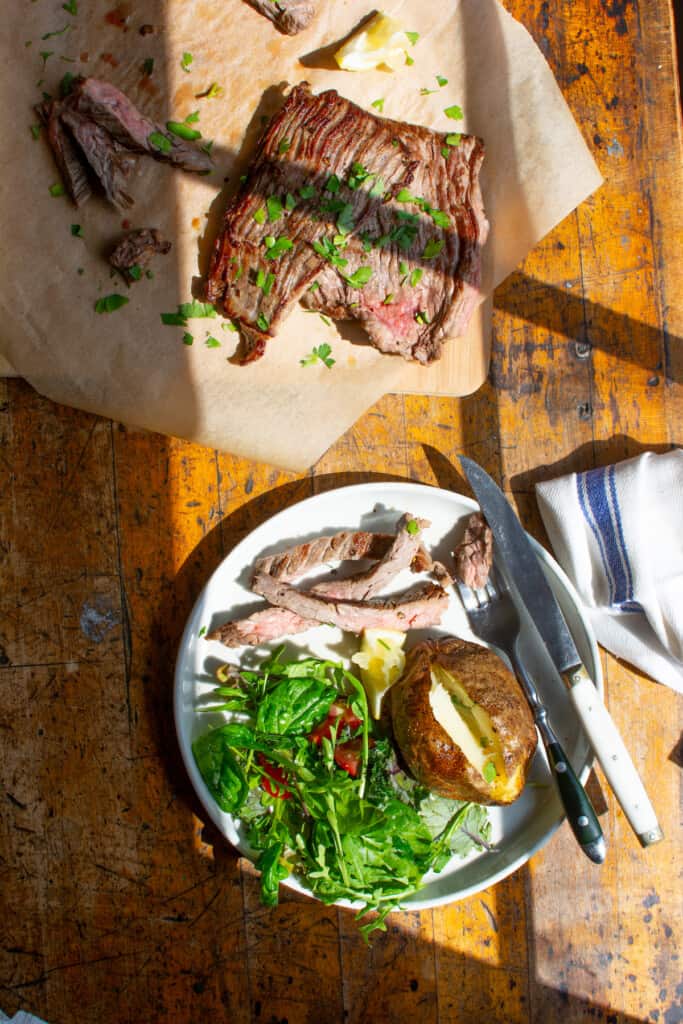 A plate with a baked potato, a salad, and several slices of skirt steak on it along with a fork and knife on a wood table. 