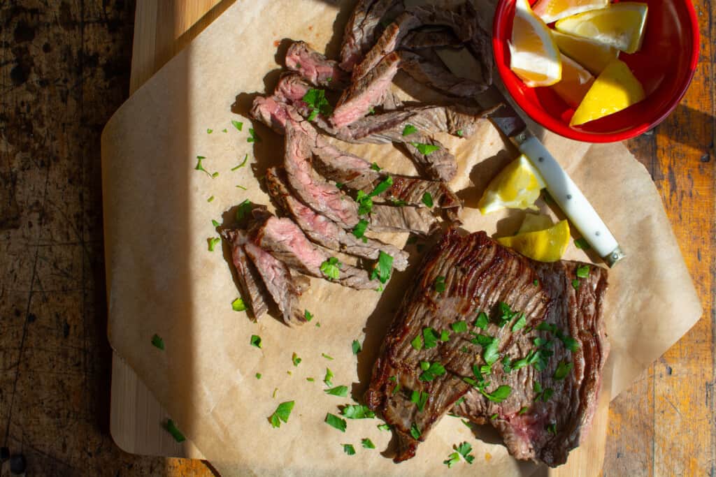 A few pieces of steak that have been cooked on a cutting board with one piece sliced into thin slices and some lemon wedges. 