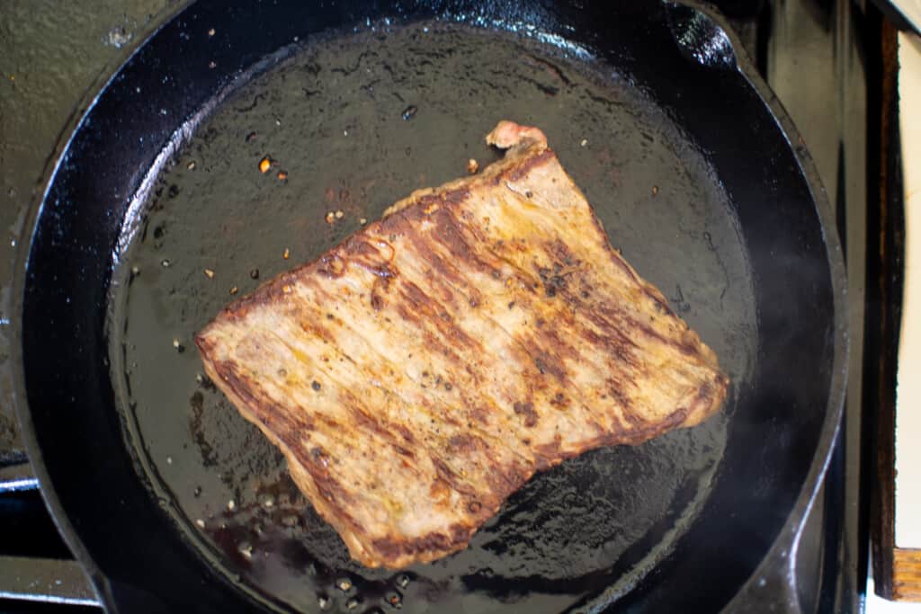 A black cast iron pan with a steak that has cooked on one side in the pan. 