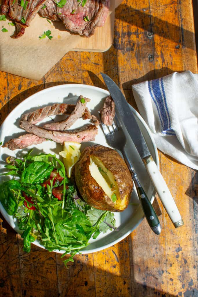 A steak dinner on a plate including a baked potato, steak, and a salad. 