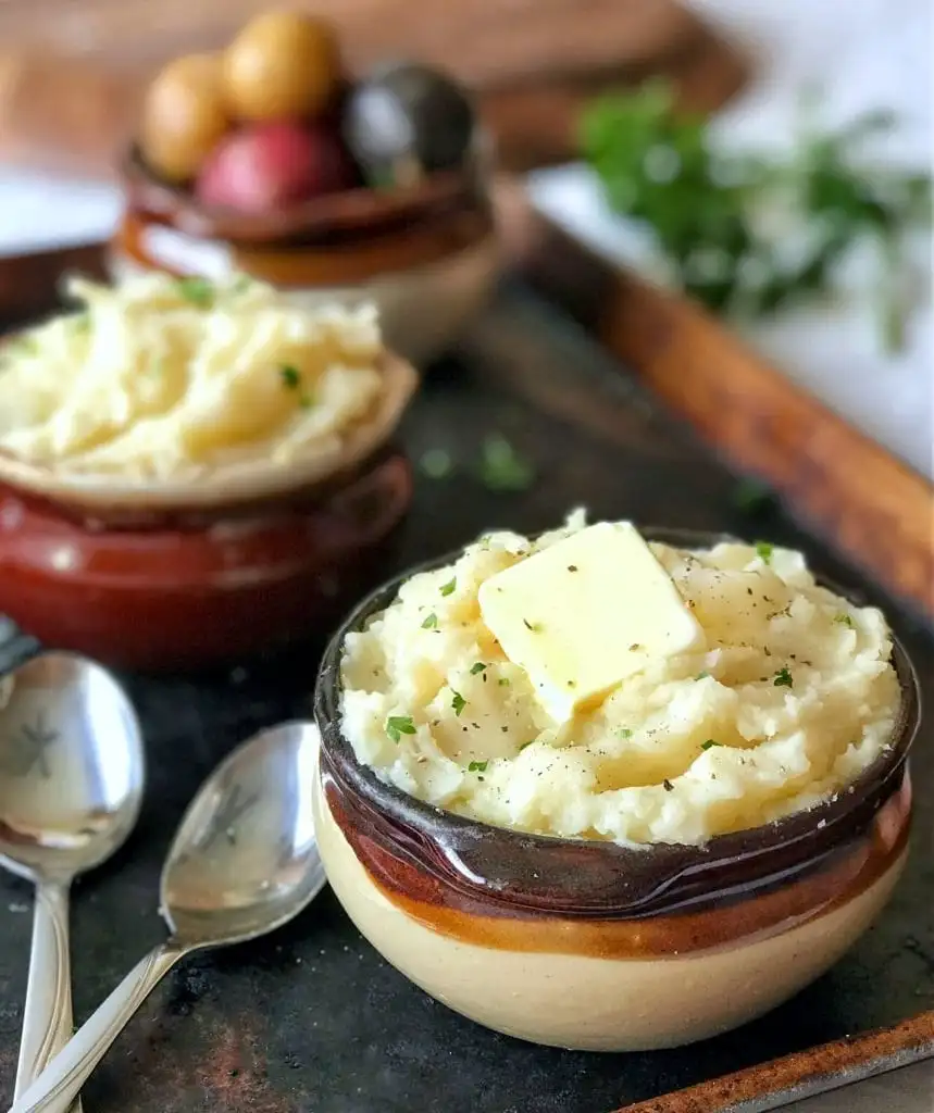 Three small ceramic bowls sitting on a baking sheet filled with mashed potatoes topped with pats of butter and spoons. 