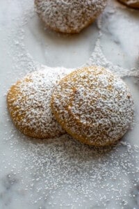 Two pumpkin cookies sprinkled with powdered sugar sitting on a marble table with another pumpkin cookie behind it.