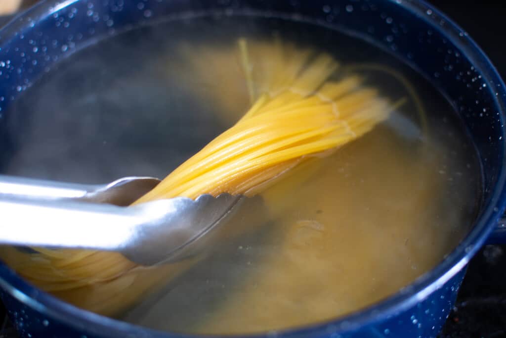 A blue pot filled with water with spaghetti in the pot being stirred by a pair of tongs. 