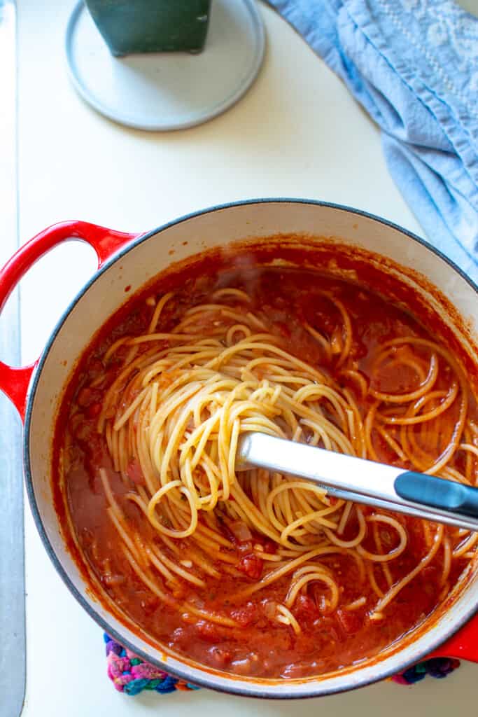 A large pot of spaghetti with marinara sauce with a pair of tongs sticking out of the sauce sitting on a white counter. 