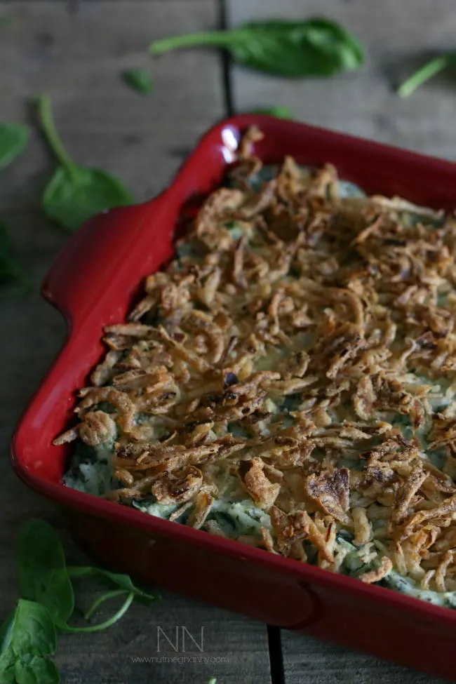 A red baking dish filled with mashed potatoes with spinach in them and topped with crispy fried onions sitting on a table. 