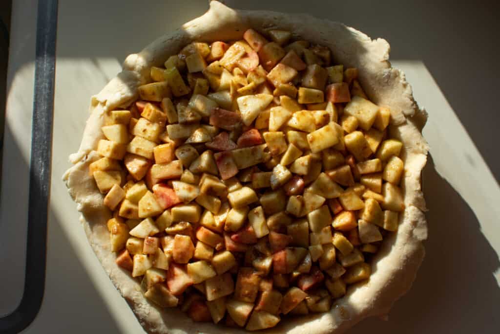 A pie being made with a bottom crust and the apples filled the pie sitting on a white counter. 