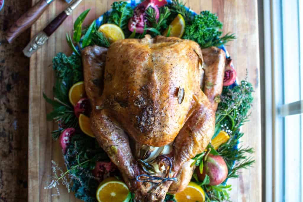 A whole, roasted turkey on a platter lined with greens and pomegranate, and orange slices. 
