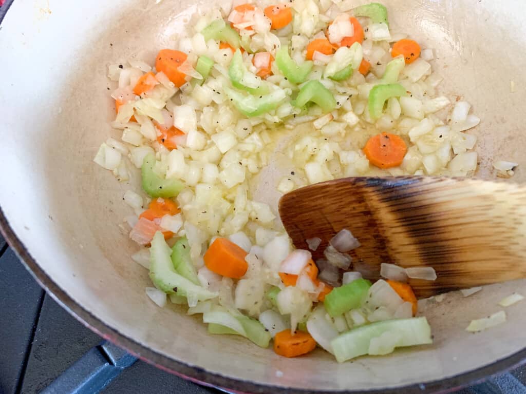 A pot of vegetables being sautéed with a wood spoon stirring the pot. 