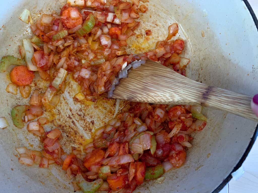 A pot of vegetables with tomato paste being sautéed in oil being stirred by a wood spoon. 