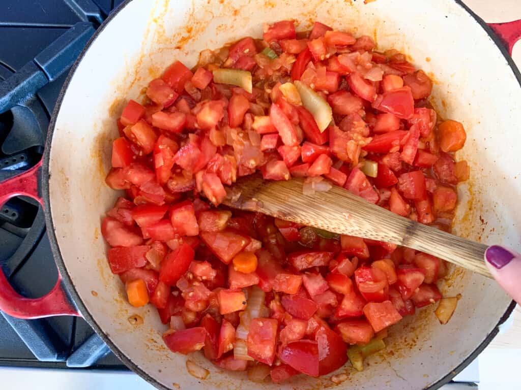 A pot on the stove with tomatoes and vegetables being stirred with a wood spoon. 