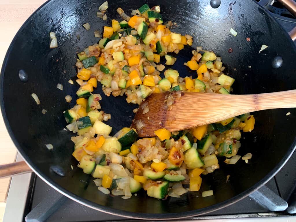 Vegetables being sautéed in a black pot on the stove with a wood spoon in the pot. 