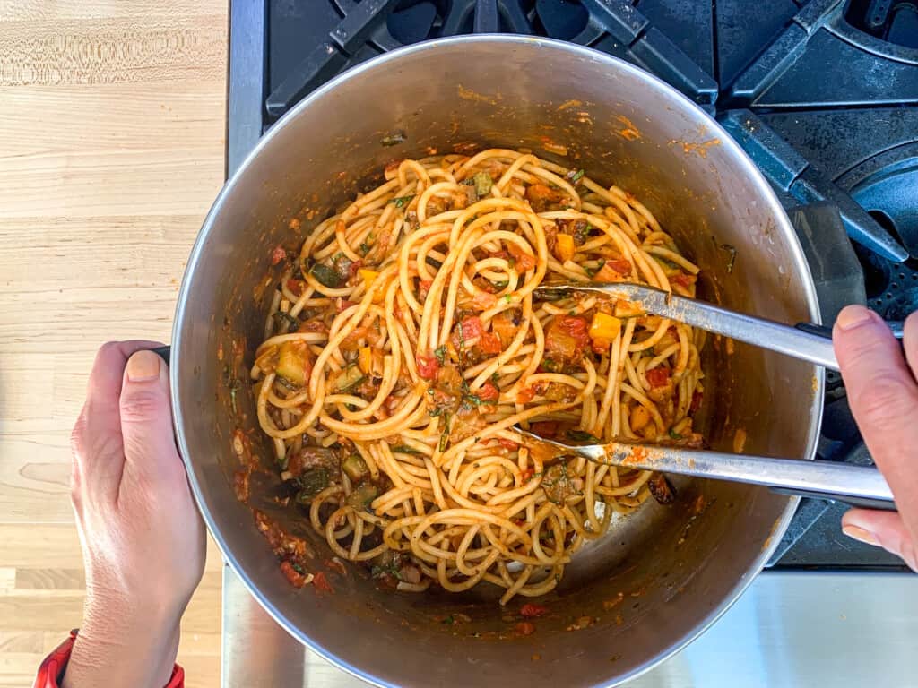 A person tossing bucatini pasta with vegan bolognese in a pot. 