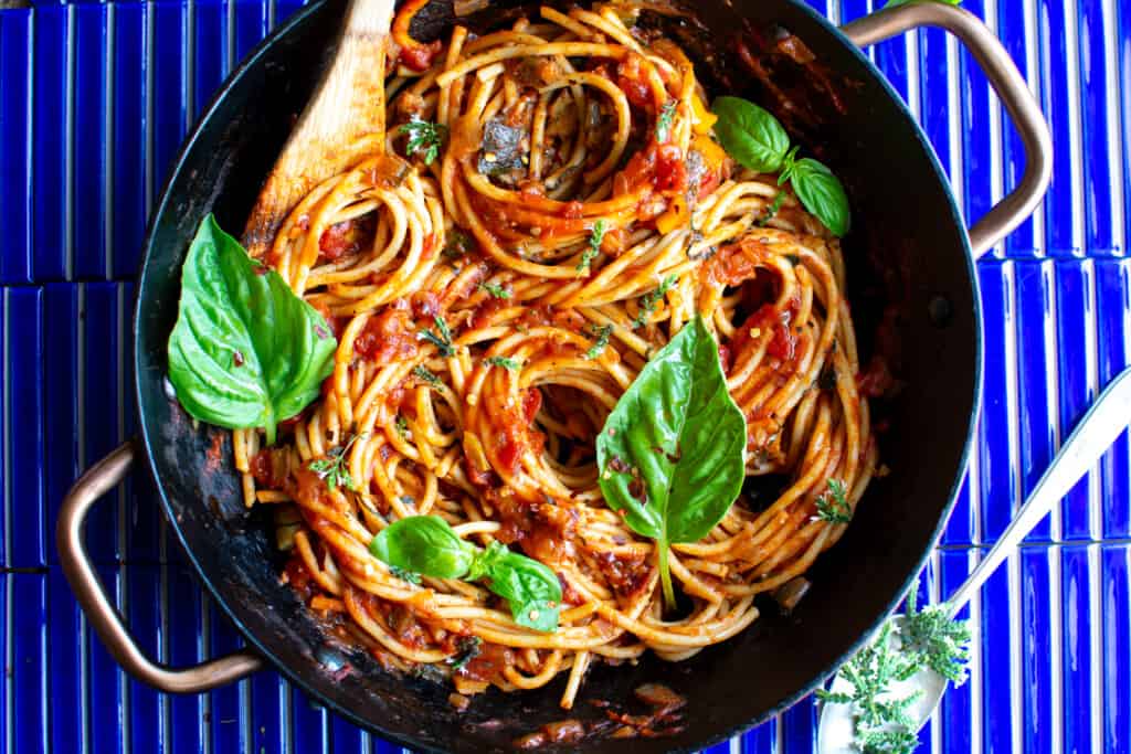 A pot of pasta with vegan bolognese sauce on a blue background with basil leaves in it. 