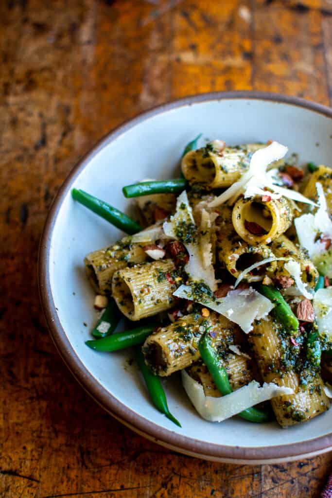 A bowl of vegan pesto pasta with shaved parmesan cheese, chopped almonds, red pepper flakes, and green beans. 