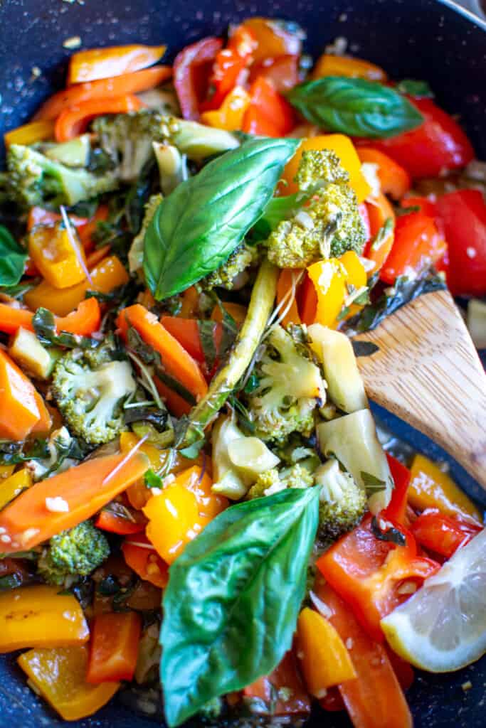 Vegetable stir fry recipe in a frying pan with basil leaves on top and a wood spoon in the pan. 