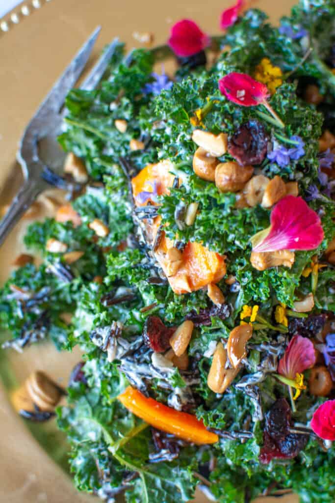 Kale salad with cashews, dried cranberries, roasted carrots, and edible flowers on a gold platter with a large fork. 